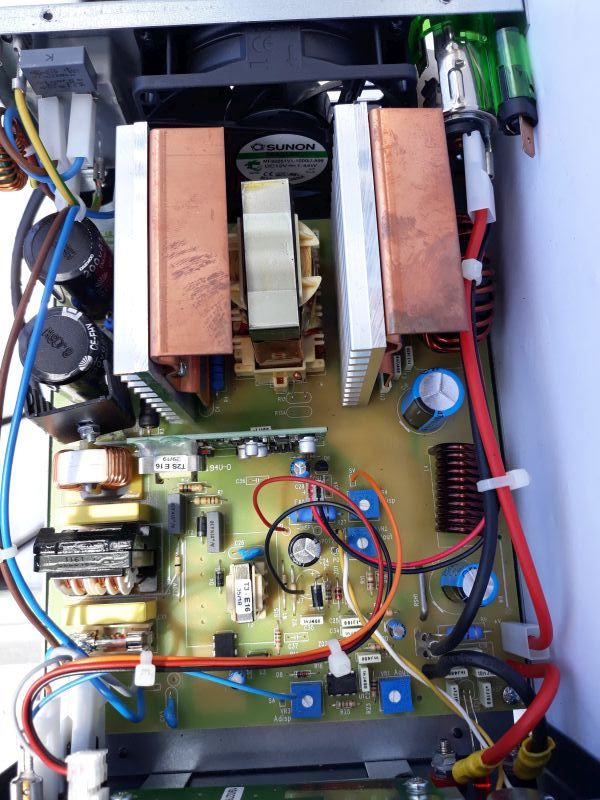 Dieser Alt-Text wurde noch nicht überprüft.

1. Kurze Zusammenfassung: Innenansicht eines geöffneten elektronischen Geräts mit bestückter Leiterplatte, großen Kühlkörpern, einem Lüfter, Spulen, Kondensatoren und mehreren farbigen Kabeln.

2. Detaillierte Beschreibung: Die Aufnahme zeigt eine gelbgrüne Leiterplatte in einem Metallgehäuse von oben. In der oberen Bildhälfte sitzt mittig ein schwarzer runder Lüfter mit sichtbarem „SUNON“-Aufkleber. Davor stehen zwei massive, kupferfarbene Blöcke auf silbernen Kühlrippen; zwischen ihnen ein beigefarbener, rechtwinkliger Transformator-Kern. Rechts oben befindet sich eine grüne Glasrohrsicherung in einer Halterung sowie dicke rote und schwarze Leitungen, mit weißen Kabelbindern am Rand fixiert. Über die Platine verteilt sind mehrere große Elektrolytkondensatoren in Blau und Schwarz, zahlreiche blaue Trimmpotentiometer, verschiedene Widerstände, Dioden und kleinere Halbleiter. Rechts unten liegt eine dunkelbraune Spule mit dicker lackierter Kupferwicklung; links sind weitere Wickelspulen und schwarze Bauteile mit Kühlblechen zu sehen. Am linken oberen Rand stehen weiße quaderförmige Komponenten, daneben verlaufen blaue und rote Leitungen. Die Leiterplatte trägt weiße Siebdruck-Beschriftungen und Markierungen; mehrere rote, gelbe, blaue und schwarze Einzeldrähte sind steckbar an den Rändern angeschlossen und kreuzen teils die Bildmitte.