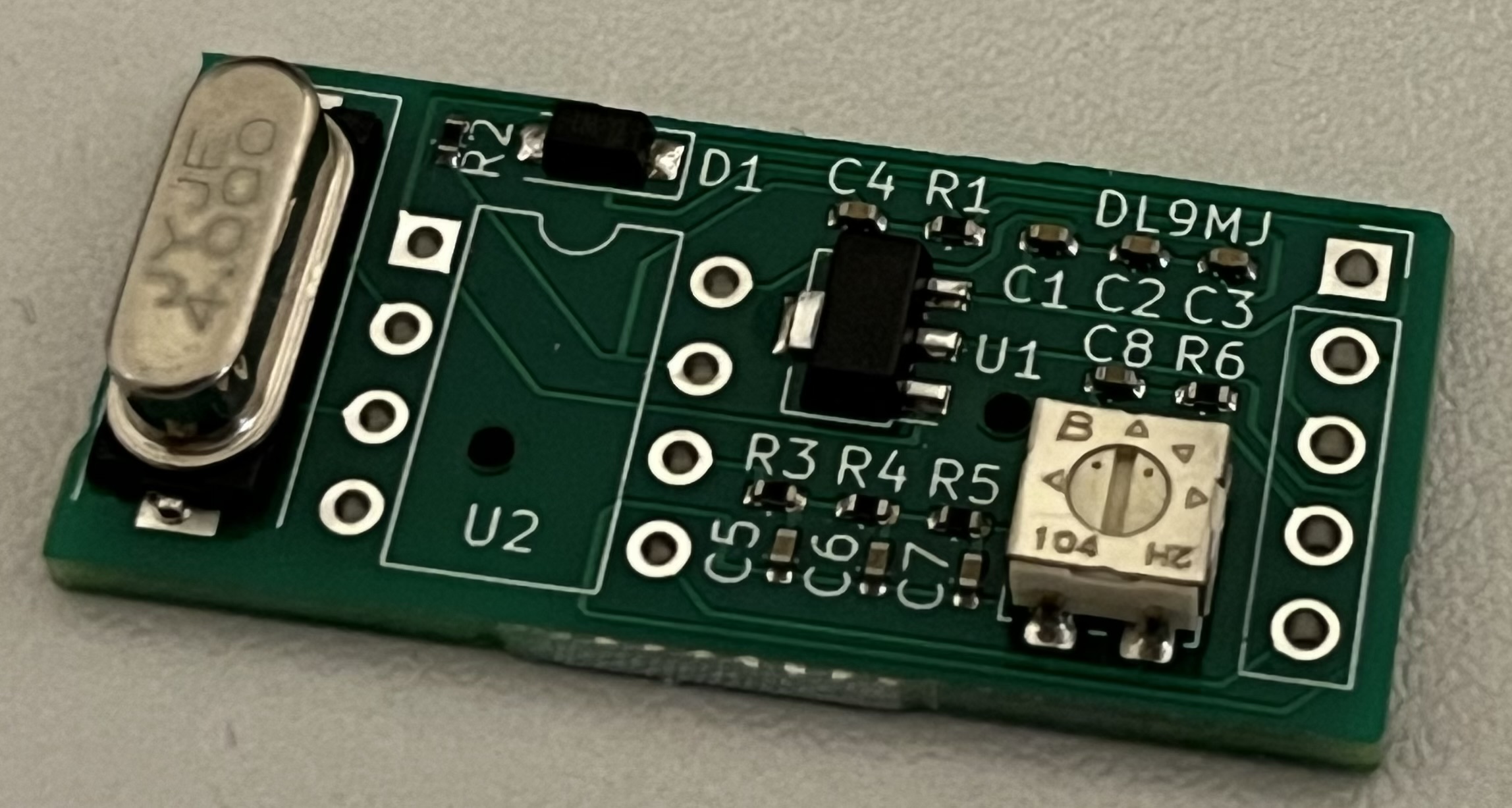 Der folgende Alt-Text wurde noch nicht geprüft: Zusammenfassung: Nahaufnahme einer kleinen grünen Leiterplatte mit mehreren SMD-Bauteilen, darunter ein metallisches Bauteil mit Aufdruck „4.000“, ein schwarzes IC-Gehäuse und ein weißes, verstellbares Trimmelement mit Schlitz.

Detaillierte Beschreibung: Links sitzt ein längliches, ovales Metallbauteil mit der Prägung „4.000“ auf einem schwarzen Sockel; daneben mehrere unbestückte, durchkontaktierte Lötaugen. Oben links ist ein kleines schwarzes Bauteil an der Position „D1“ zu sehen, umlaufend sind mit weißem Bestückungsdruck diverse Bezeichnungen markiert, u. a. „R2“, „C4“, „R1“, „C1“, „C2“, „C3“, „U1“, „C8“, „R6“, „R3“, „R4“, „R5“, „U2“ sowie „DL9MJ“. Rechts der Mitte befindet sich ein schwarzes SMD-Bauteil mit mehreren Anschlüssen, darunter weitere kleine Widerstände und Kondensatoren. Rechts unten ist ein weißes, quadratisches Trimmelement mit Schlitz zur Justage erkennbar, mit Aufdrucken „B“ und „104“; seine Lötfahnen ragen nach unten. Entlang der rechten Kante befinden sich weitere Reihen unbestückter, durchkontaktierter Lötaugen. Die Leiterplatte liegt auf einer hellgrauen, leicht strukturierten Oberfläche.