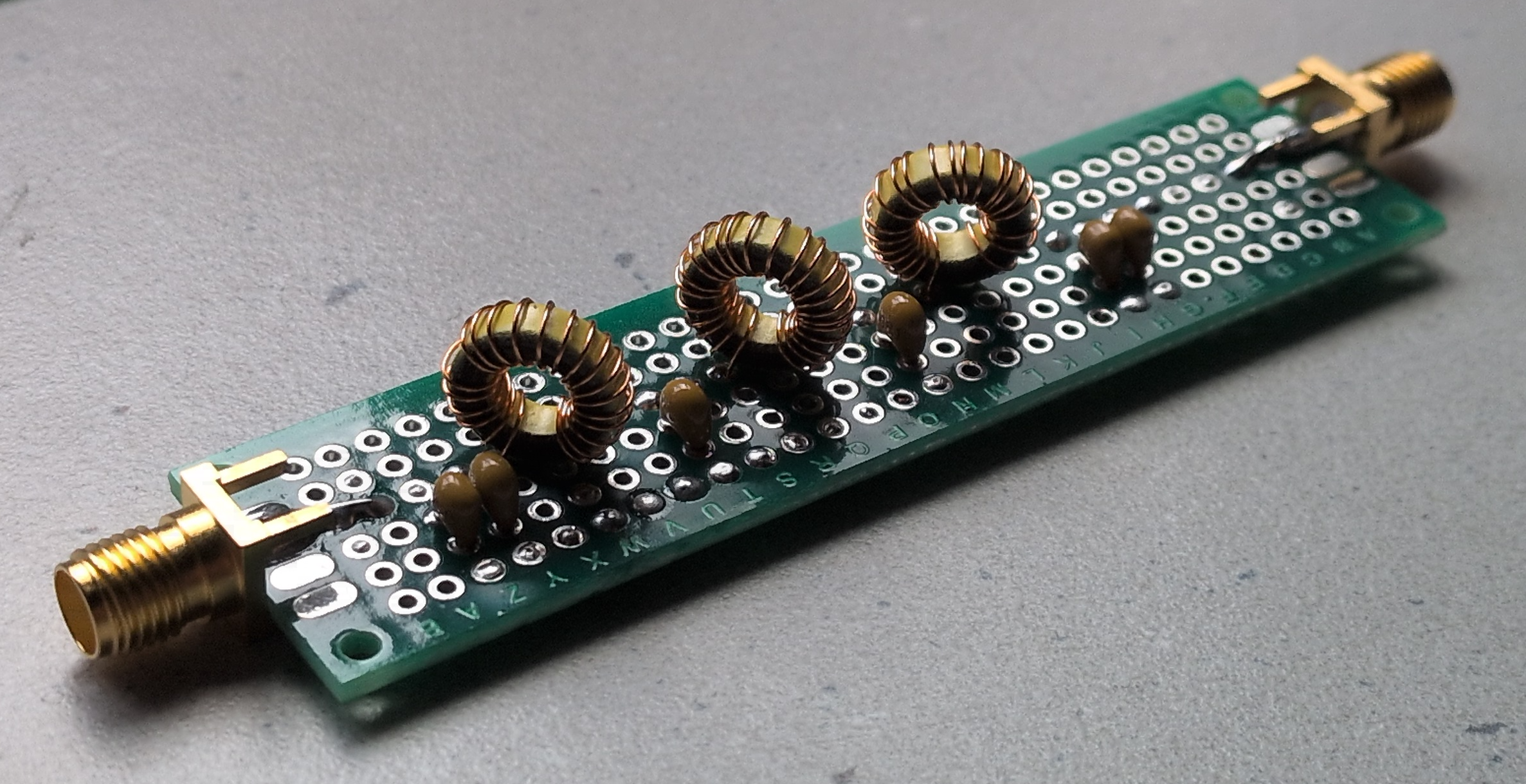 Dieser Alt-Text wurde noch nicht überprüft.

1. Zusammenfassung: Langer, schmaler Elektronikaufbau auf grünem Lochrasterboard mit drei ringförmigen Kupferspulen und mehreren kleinen braunen Bauteilen zwischen zwei goldfarbenen Koaxanschlüssen.

2. Detaillierte Beschreibung: Auf einem rechteckigen, grünen Lochrasterboard mit Reihen aus runden Lötaugen sind drei gleichartige, ringförmige Spulen aus lackiertem Kupferdraht auf gelblichen Kernen in einer Linie angeordnet. Dazwischen und daneben sitzen mehrere kleine, braun glänzende, tropfenförmige Bauteile (per Drahtanschluss eingelötet). An beiden Stirnseiten des Boards befinden sich goldfarbene, koaxiale Buchsen/Stecker mit verlöteten Mittelkontakten und seitlichen Lötfahnen zur Masse. Die Lötstellen sind sichtbar, teils mit dunklen Flussmittelspuren. Das Board liegt schräg auf einer grauen, leicht strukturierten Oberfläche; die Beleuchtung kommt von oben links und erzeugt weiche Schatten.
