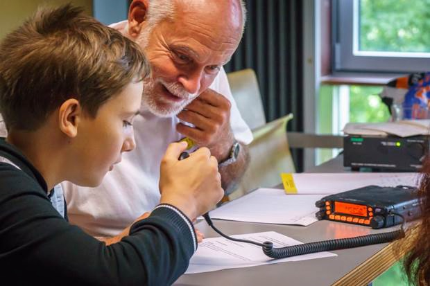 Der folgende Alt-Text wurde noch nicht geprüft: 1) Kurzbeschreibung: Ein Junge sitzt mit einem älteren Mann an einem Tisch und spricht in ein Handmikrofon, das über ein Spiralkabel mit einem Funkgerät mit orange beleuchtetem Display verbunden ist.

2) Detaillierte Beschreibung: Links im Bild hält der Junge das Handmikrofon nah an den Mund, während der ältere Mann neben ihm leicht nach vorn gebeugt zuschaut und eine Hand am Kinn hat. Auf dem Tisch liegen mehrere Blätter Papier mit Textzeilen sowie ein Stift; das Spiralkabel des Mikrofons führt zu einem kompakten, schwarzen Funkgerät mit orange beleuchtetem Display und mehreren Tasten auf der rechten Seite des Tisches. Im Hintergrund stehen weitere elektronische Geräte, darunter ein dunkles Gerät mit grünem Display. Der Raum ist hell, durch ein geöffnetes Fenster fällt Tageslicht; draußen sind grüne Baumkronen zu sehen.