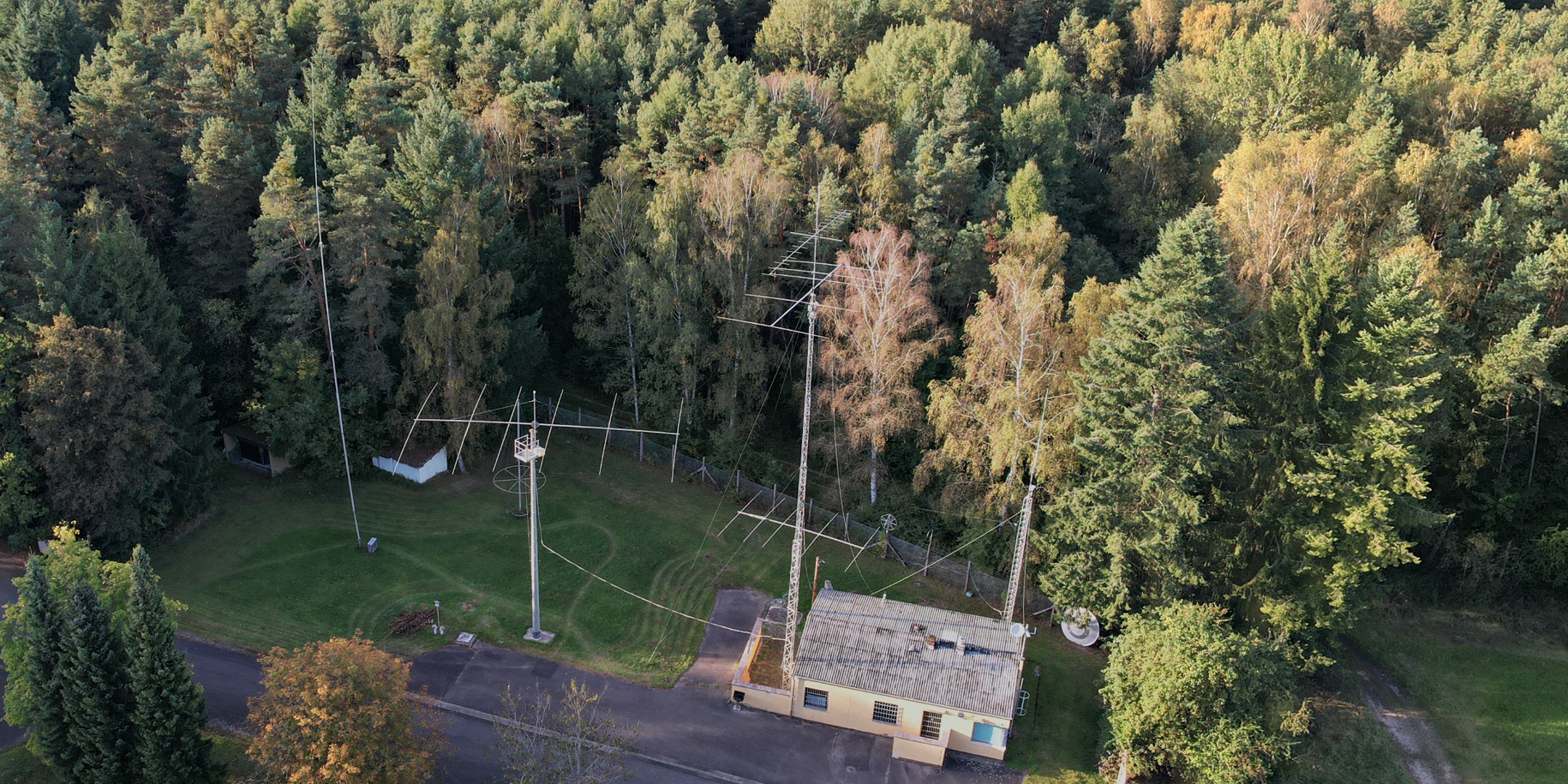 Der folgende Alt-Text wurde noch nicht geprüft: 1) Zusammenfassung: Luftaufnahme eines Gebäudes am Waldrand mit mehreren hohen Antennenmasten und großflächigen Richtantennen auf einer Wiese.

2) Details: Rechts neben dem flachen, cremefarbenen Gebäude mit Wellblechdach, Fenstern und kleiner Satellitenschüssel an der Ecke stehen zwei hohe Gittermasten, jeweils mit mehreren horizontalen Antennenträgern und zahlreichen querstehenden Elementen; die Masten sind mit Abspannseilen gesichert, zwischen ihnen verlaufen gespannte Leitungen. In der Platzmitte steht ein Betonmast mit runder Arbeitsplattform und Leiter. Links befindet sich ein sehr schlanker, hoher Mast nahe einem kleinen offenen Unterstand. Der Bereich ist eine gemähte Rasenfläche mit sichtbaren Mähspuren, teils von einem Zaun begrenzt, und wird von dichtem Mischwald mit grünen bis gelblichen Baumkronen umgeben; unten im Bild verläuft eine schmale Asphaltstraße.
