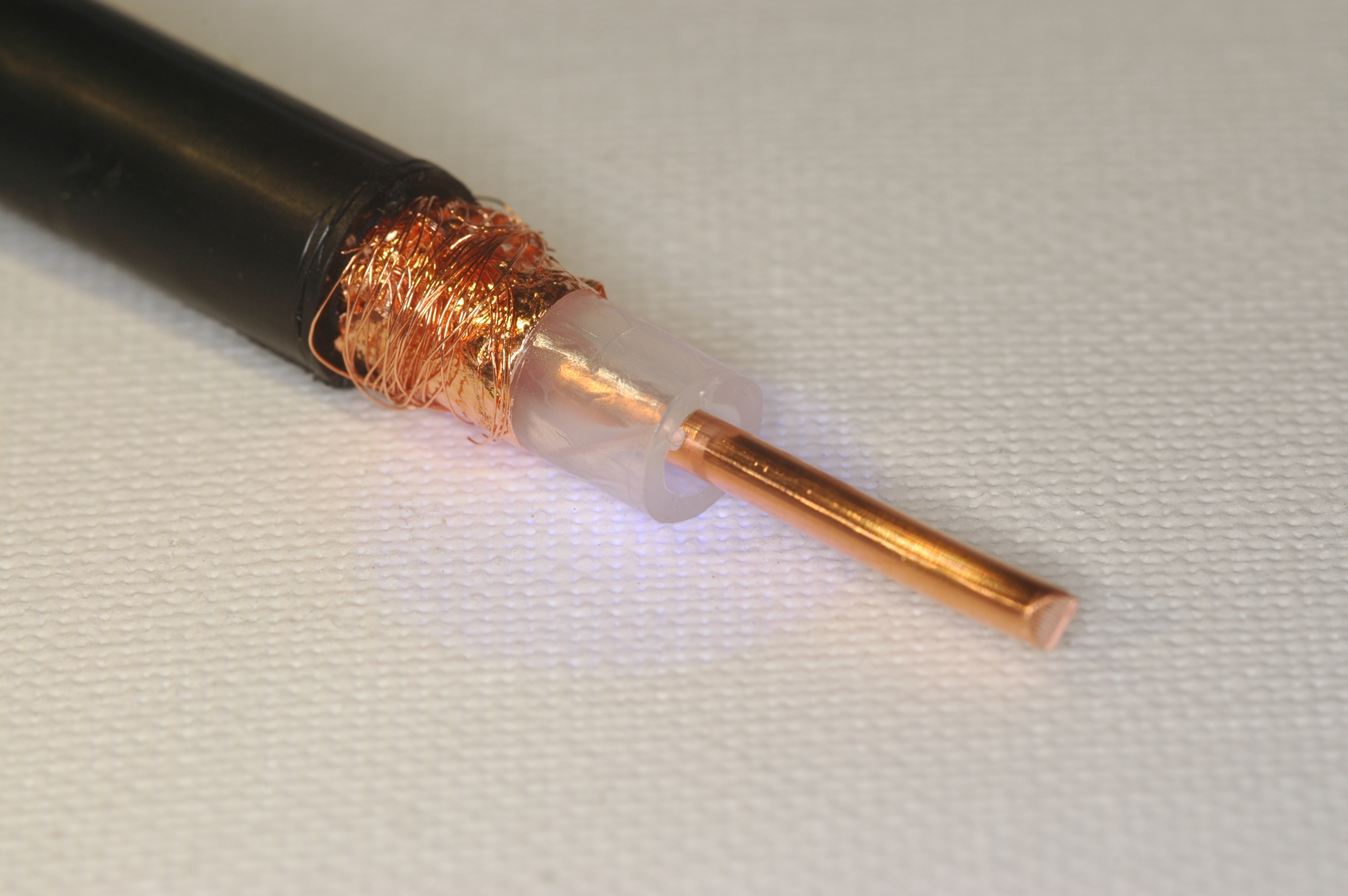 Der folgende Alt-Text wurde noch nicht geprüft: 1) Kurze Zusammenfassung: Nahaufnahme eines abisolierten Koaxialkabels mit sichtbarem Kupfer-Innenleiter, weißem Isolator und freigelegter Kupferabschirmung.

2) Detaillierte Beschreibung: Das Kabel liegt schräg auf einer weißen, fein strukturierten Unterlage. Links ist der schwarze Außenmantel zu sehen, an dessen Ende blanke, glänzende Kupferlitzen und -folie freiliegen und teilweise als dünne Drähte um das Kabel gewickelt sind. Daran schließt sich ein kurzes, transluzentes, weißliches Kunststoffröhrchen an, in dessen Mitte der gerade, massige, rötlich glänzende Kupfer-Innenleiter nach rechts hervorsteht. Das Foto ist stark vergrößert, mit weichen Lichtreflexen auf Metall und Kunststoff.