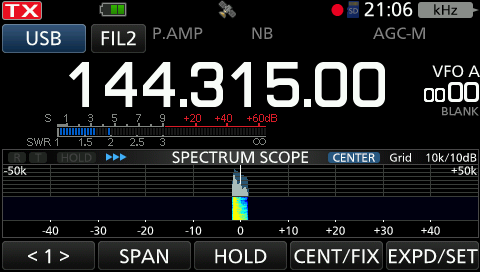 Der folgende Alt-Text wurde noch nicht geprüft: 1) Kurze Zusammenfassung: Anzeige eines Funkgerätedisplays im USB-Betrieb bei 144.315.00 mit roter TX-Anzeige und einem Spektrums-Peak in der Mitte.

2) Detaillierte Beschreibung: Oben links leuchtet ein rotes „TX“-Feld, daneben sind Schaltflächen/Indikatoren „USB“ und „FIL2“ sowie kleine Symbole (u. a. ein grünes Batteriesymbol und ein Satellitensymbol) zu sehen; außerdem „P.AMP“, „NB“ und „AGC-M“. Rechts oben steht die Uhrzeit „21:06“ und „kHz“, daneben ein roter Punkt. Zentral dominiert die große Frequenzanzeige „144.315.00“. Rechts davon steht „VFO A“ und darunter eine Nullanzeige sowie der Schriftzug „BLANK“. Oberhalb der Mitte befindet sich eine S‑Skala mit Markierungen „S“ und dB‑Werten (−20, +20, +40, +60 dB); darunter eine SWR‑Skala („SWR 1“). Im unteren Bildschirmbereich zeigt der „SPECTRUM SCOPE“ (Modus „CENTER“, Grid „10k/10dB“) einen horizontalen Frequenzbereich von −50k bis +50k mit vertikalen Amplitudenmarken (−40 bis +40); in der Mitte bei 0 ist ein schmaler, farbiger Ausschlag in Blau/Grün/Gelb sichtbar. Ganz unten sind Softkeys mit den Beschriftungen „<1“, „SPAN“, „HOLD“, „CENT/FIX“ und „EXPD/SET“ eingeblendet.