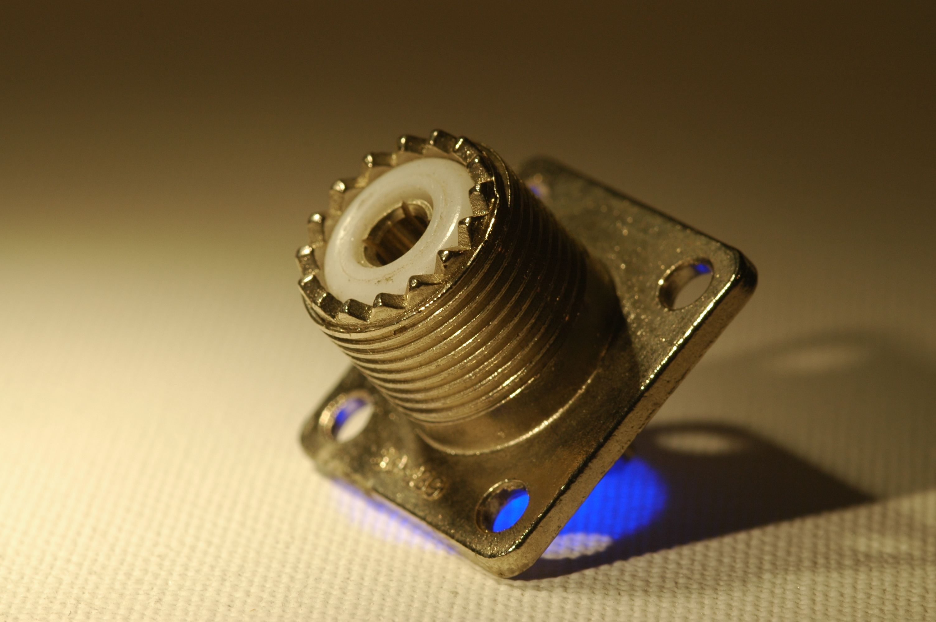 1) Kurzbeschreibung: Koaxiale PL-Einbaubuchse mit Außengewinde und weißem Isolator.

2) Ausführliche Beschreibung: Das Foto zeigt eine koaxiale PL-Einbaubuchse mit Außengewinde und weißem Isolator um eine Öffnung in der Mitte.