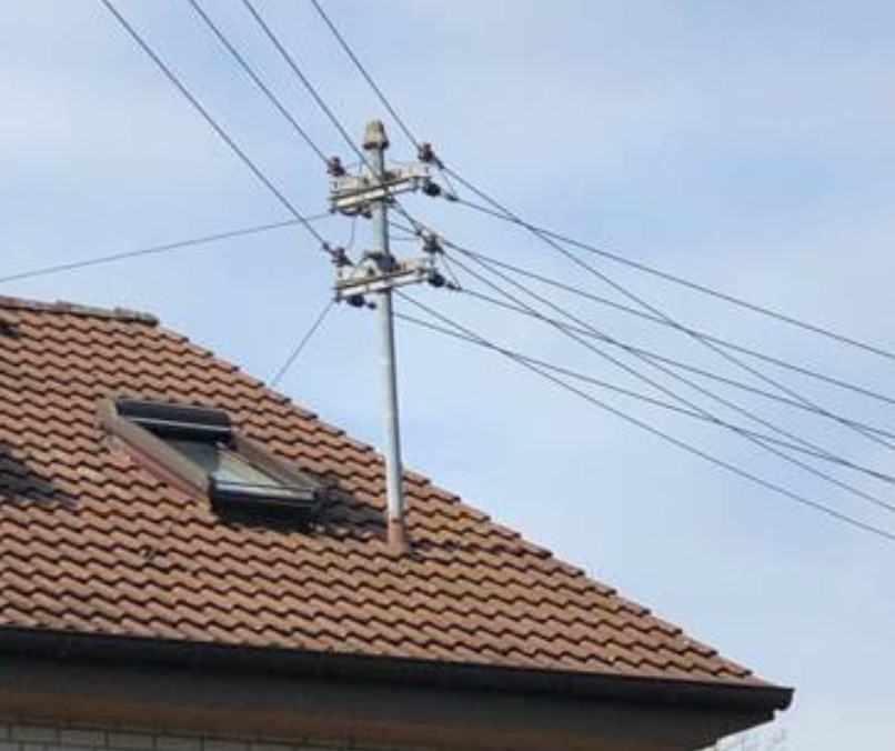 Der folgende Alt-Text wurde noch nicht geprüft: Zusammenfassung: Schräges Ziegeldach mit Dachfenster und einem auf dem Dach montierten Freileitungsmast, an dem mehrere Leitungen vorbeiführen, vor hellem Himmel.

Details: Zu sehen ist der obere Teil eines Hauses mit rotbraunen Dachziegeln; links befindet sich ein rechteckiges Dachfenster mit dunklem Rahmen. Aus dem Dach ragt ein metallischer Mast mit zwei Querträgern, auf denen mehrere Isolatoren montiert sind. Mehrere gespannte Leitungen verlaufen schräg quer durchs Bild von rechts nach links. Am unteren Rand ist die Dachkante sichtbar, der Hintergrund besteht aus einem hellblauen, leicht bewölkten Himmel.