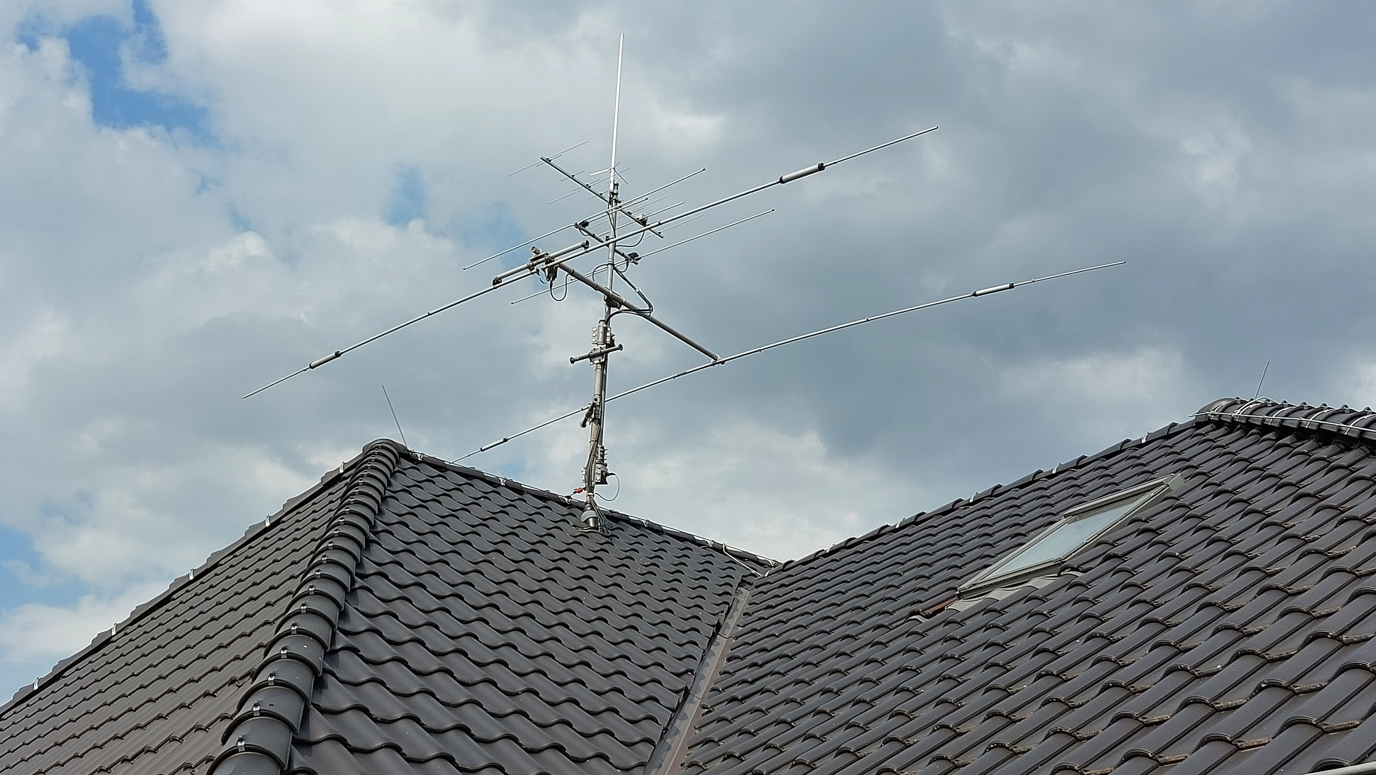 1) Kurzbeschreibung: Metallmast auf einem Dach mit mehreren Antennen.

2) Ausführliche Beschreibung: Das Foto zeigt einen Antennenmast, der auf einem Hausdach installiert ist. Unten befindet sich eine große Kurzwellen-Richtantenne mit zwei Elementen, darüber eine kleinere 2-Element-Antenne für VHF und darüber eine UHF-Richtantenne mit mehreren Elementen. An der Spitze ist eine vertikale Rundstrahlantenne für UKW montiert.