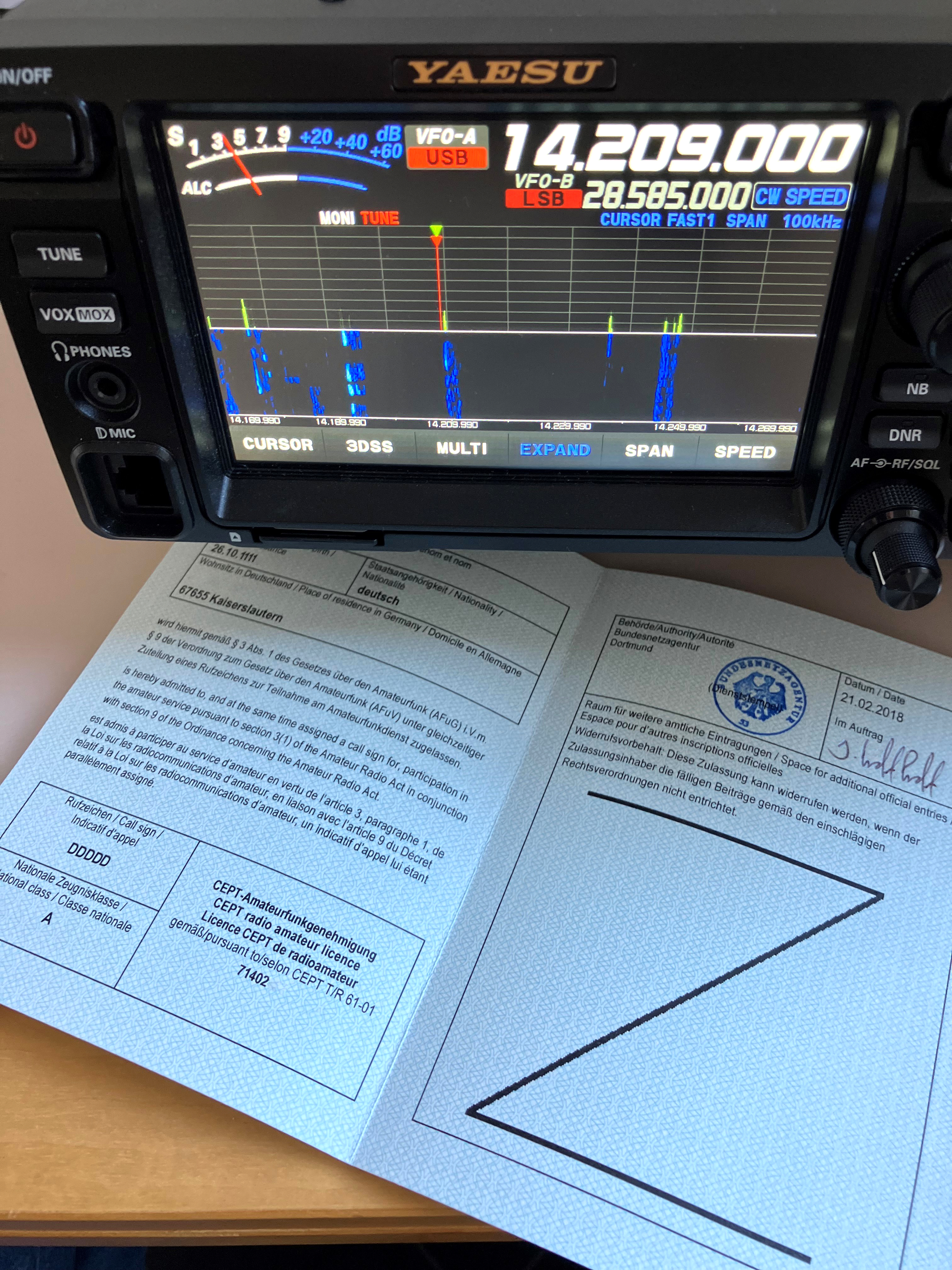 Der folgende Alt-Text wurde noch nicht geprüft: Kurzfassung: Ein Yaesu-Kurzwellen-Transceiver mit aktivem Spektrumdisplay steht auf einem Schreibtisch, davor liegt ein geöffnetes, blaues CEPT-Amateurfunkdokument mit Stempel und Unterschrift.

Detaillierte Beschreibung: Im oberen Bildbereich dominiert die Front eines YAESU-Funkgeräts; auf dem farbigen Display sind rechts groß die Frequenz 14.209.000 und die Betriebsart USB (VFO-A) eingeblendet, darunter VFO-B mit 28.585.000 und LSB sowie die Angabe SPAN 100 kHz. Ein Spektrum-/Wasserfalldiagramm zeigt mehrere schmale, vertikale Signalspuren in Blau und Grün vor dunklem Hintergrund; am unteren Displayrand ist eine Frequenzskala um 14,208–14,209 MHz erkennbar. Links oben befindet sich eine S-Meter/ALC-Anzeige (S1 bis S9, +20 bis +60 dB). Unter dem Display stehen Tastenbezeichnungen wie CURSOR, 3DSS, MULTI, EXPAND, SPAN und SPEED. Links am Gerät sind Tasten mit Aufdruck TUNE, VOX/MOX und PHONES sowie eine Mikrofonbuchse zu sehen; rechts befinden sich Tasten/Regler mit Bezeichnungen NB, DNR und AF-G/RF/SQL. Vor dem Gerät liegt auf einer Holzoberfläche ein aufgeklapptes, hellblau gemustertes CEPT-Amateurfunkdokument mit mehrsprachigem Text; auf der rechten Seite ist ein runder Amtsstempel mit Datum 21.02.2018 und Ortsangabe Dortmund sowie eine Unterschrift sichtbar, darunter ein großes, zickzackförmiges Linienfeld.
