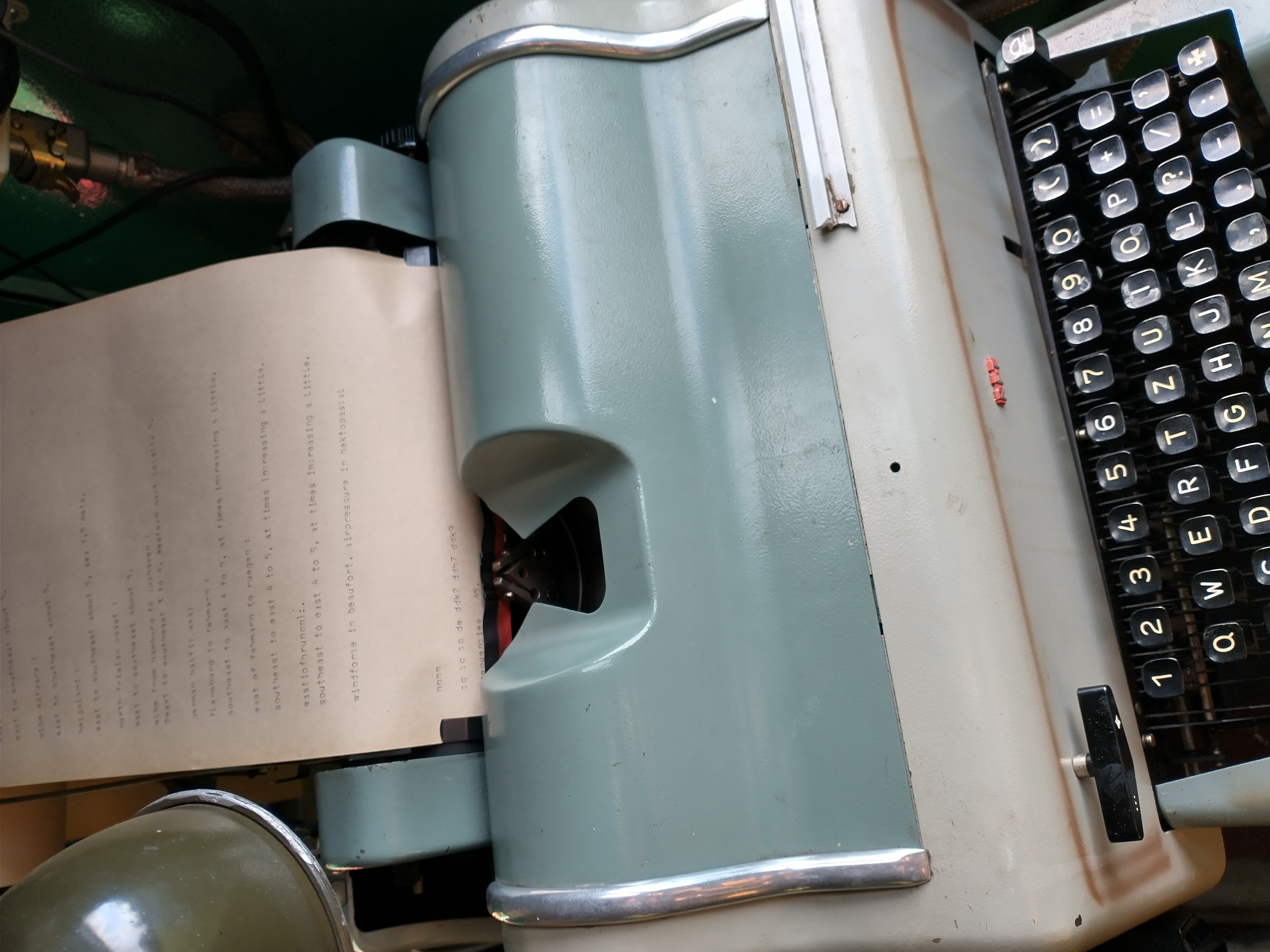 1) Kurzbeschreibung: Mintgrün-beiger Fernschreiber mit eingelegtem Papier.

2) Ausführliche Beschreibung: Auf dem Foto ist ein an den Kanten abgerundetes Gerät in Mintgrün und Beige mit polierten Metallleisten links und rechts und einer Papierführung oben zu sehen. Ein cremefarbenes Blatt ist eingespannt. Unten befindet sich eine schwarze Tastatur mit weiß beschrifteten, leicht gewölbten Tasten: oben die Zahlenreihe 1–0, darunter ein QWERTZ-Layout mit deutschen Umlauten.